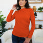 Woman modeling an orange FAM-FAM Ruched Mock Neck Long Sleeve T-Shirt, perfect for fall layering and style, in a cozy room setting.