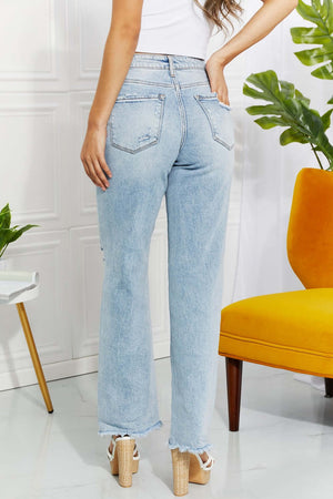 Back view of woman wearing high waist light wash Allie 90's Dad Jean with distressed detailing and raw hem, standing near yellow chair.