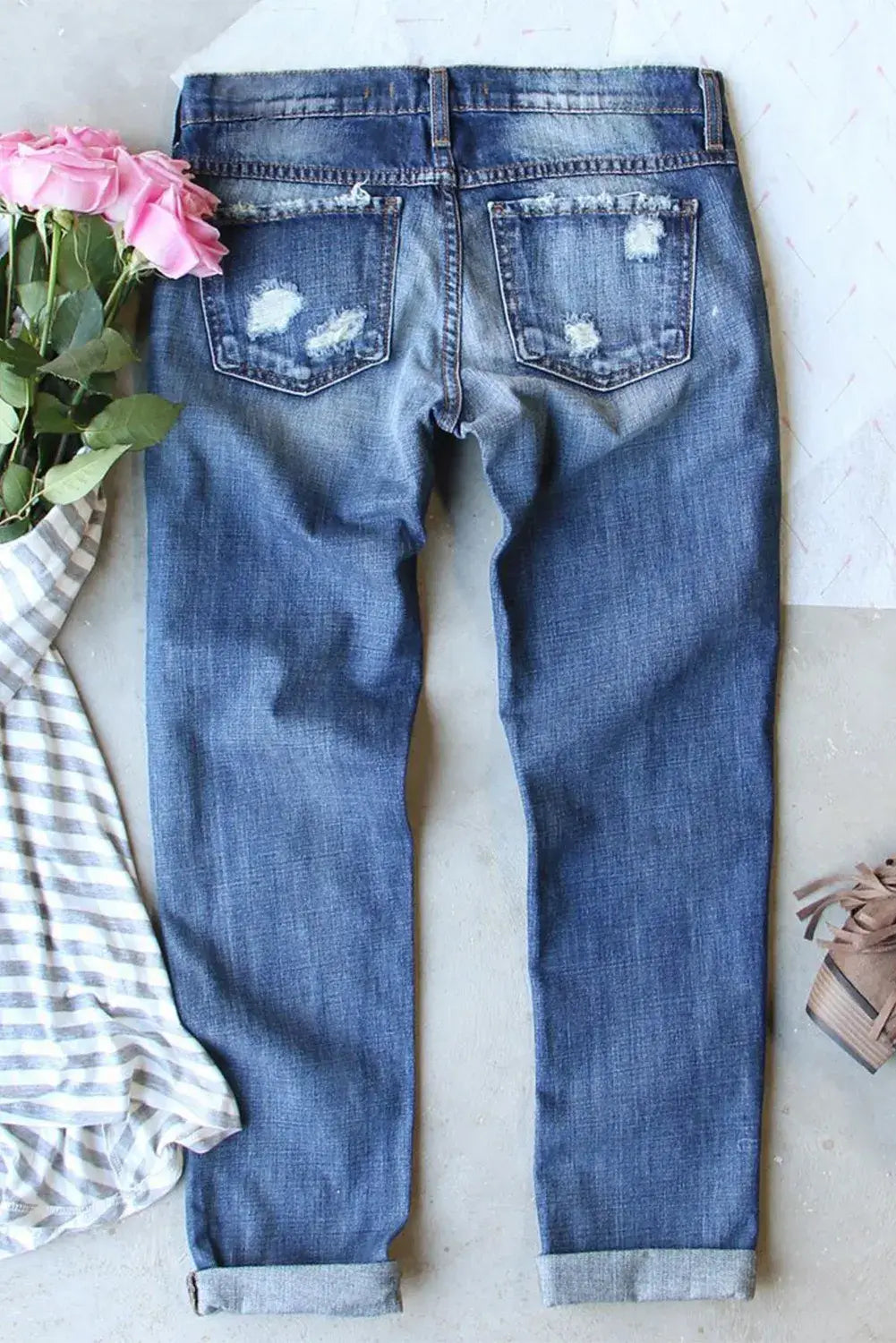 Back view of distressed skeleton pattern jeans with pockets, slightly stretchy denim, laid flat with a striped shirt, pink roses, and brown shoes nearby.