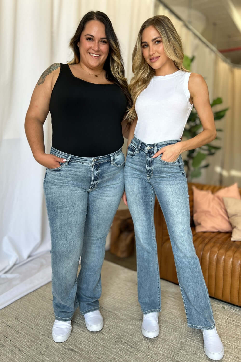 Two women wearing Judy Blue medium rise bootcut jeans pose indoors, showcasing denim's flattering fit with casual tops and sneakers.