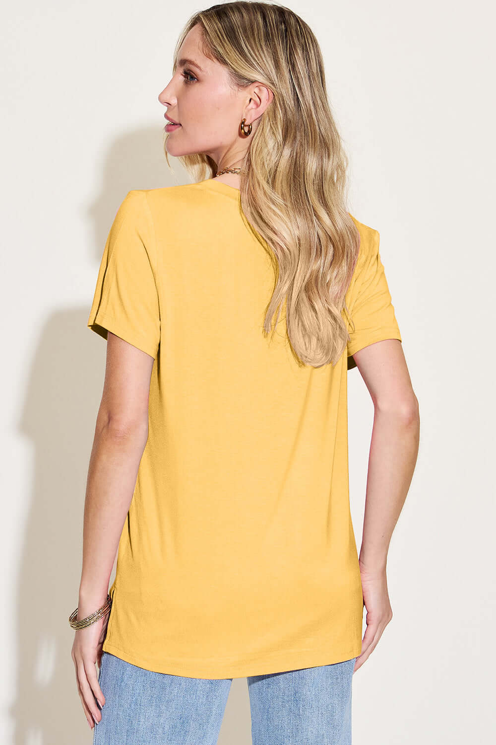 Woman wearing a yellow Bamboo V-Neck High-Low T-Shirt, showing back view, paired with blue jeans, highlighting comfortable fit and slight stretch.