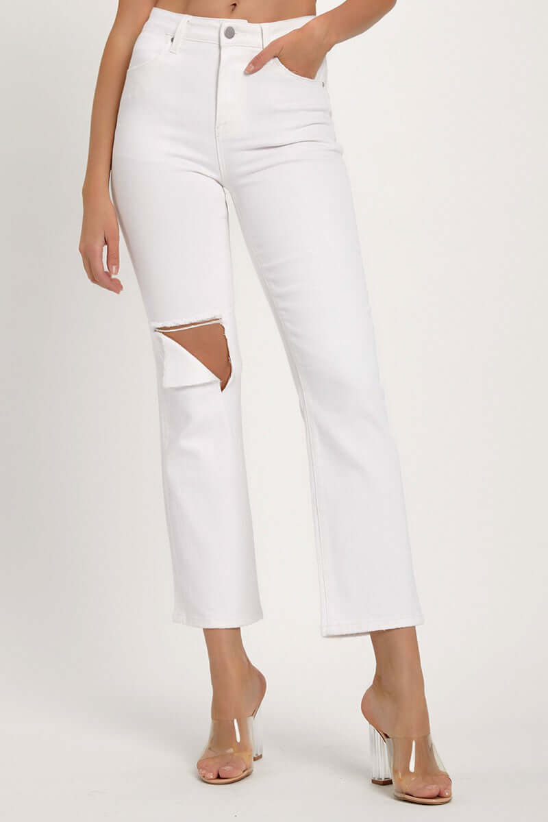 Woman modeling RISEN distressed cropped white jeans with a straight leg cut and edgy knee rip, paired with clear heels.