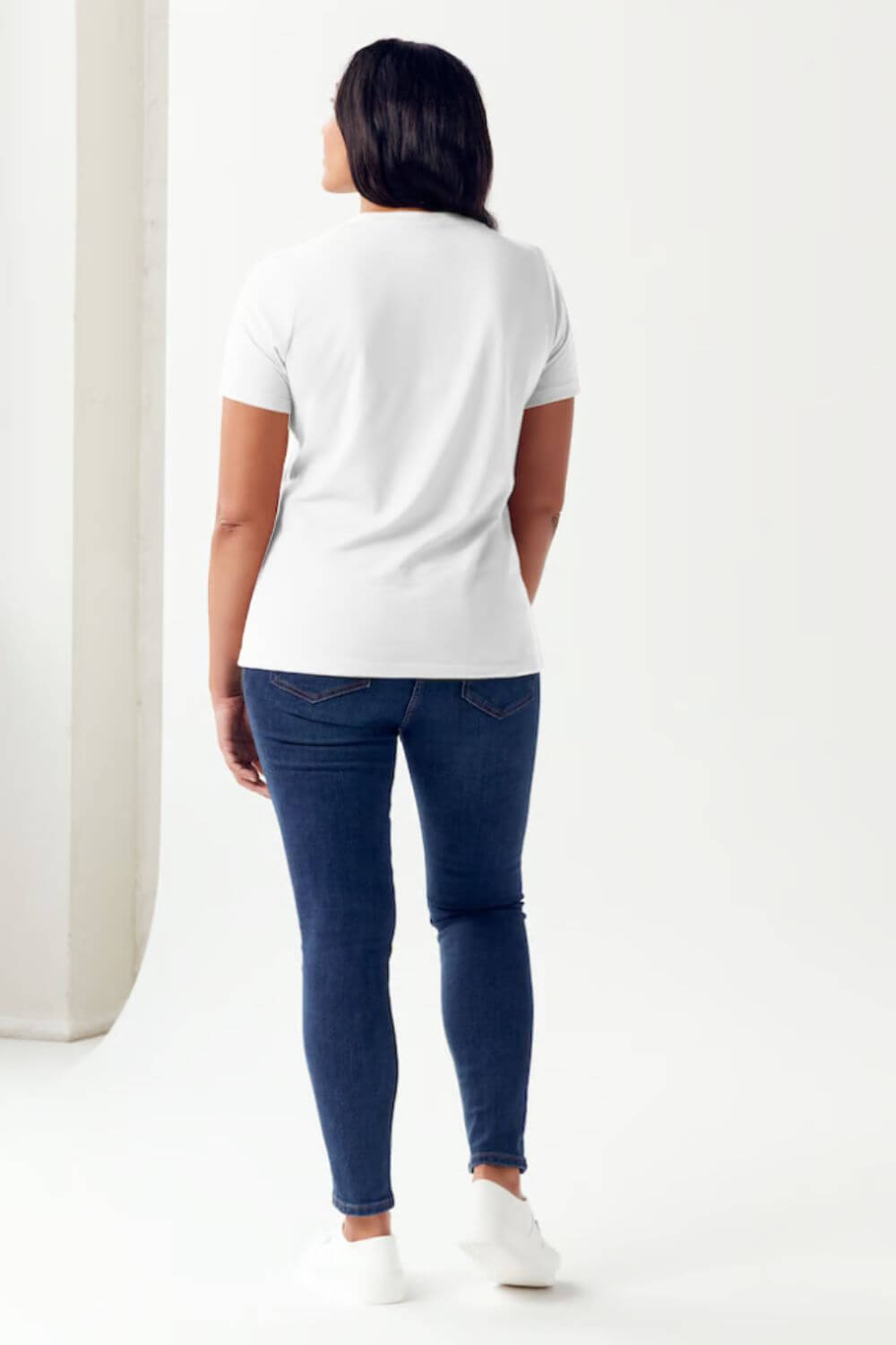 Woman wearing a white round neck short sleeve t-shirt and blue jeans, standing with her back to the camera.