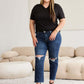 Plus-size woman wearing RFM Jeans Tummy Control Distressed High Waist Raw Hem Jeans, standing in a stylish modern living room.