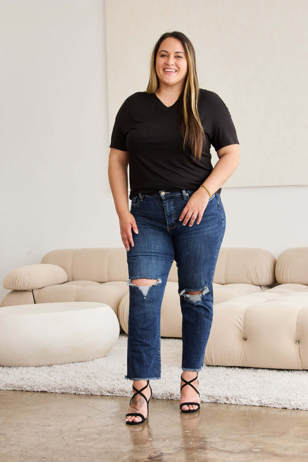 Plus-size woman wearing RFM Jeans Tummy Control Distressed High Waist Raw Hem Jeans, standing in a stylish modern living room.