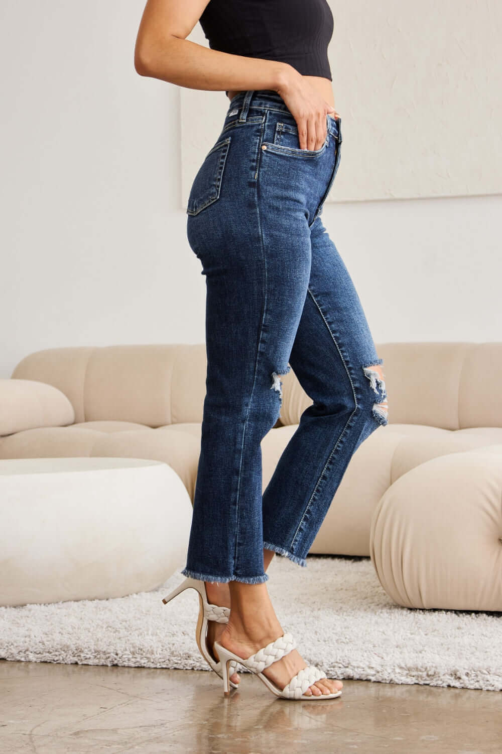 Woman wearing RFM Jeans Crop Dylan Full Size Tummy Control Distressed High Waist Raw Hem Jeans with white heels in a stylish room