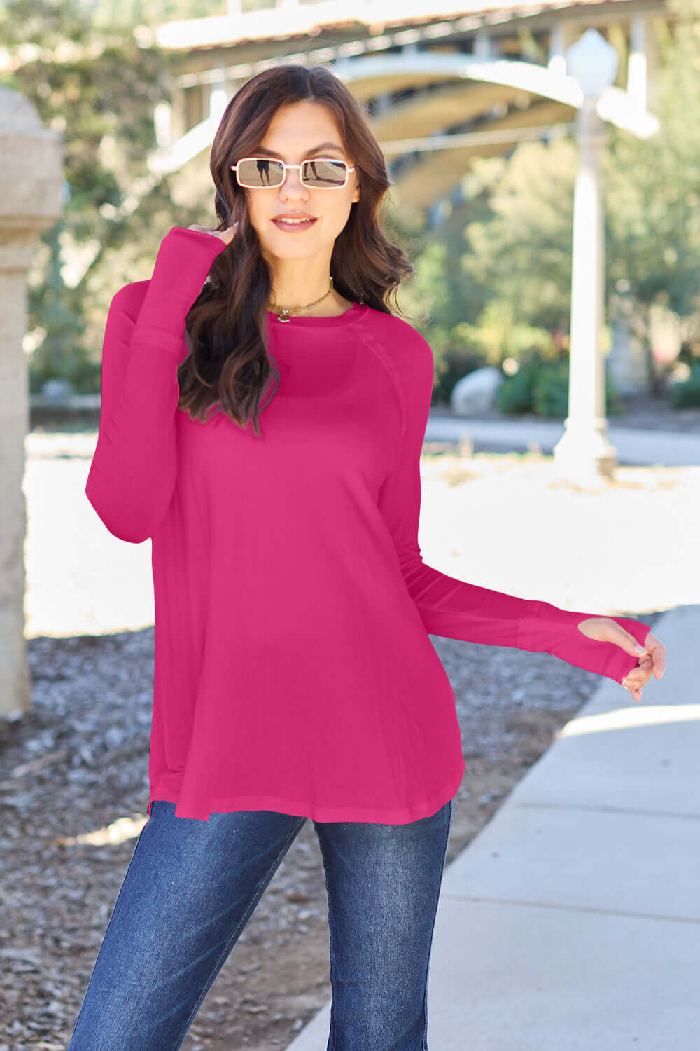 Woman wearing a round neck long sleeve t-shirt in vibrant pink with blue jeans, posing outdoors on a sunny day.