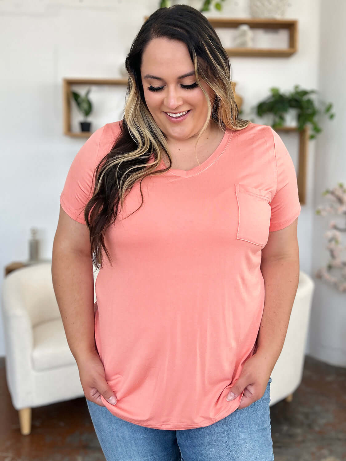 Woman wearing Basic Bae Bamboo Full Size V-Neck Short Sleeve T-Shirt in peach color, smiling and standing in stylish living room.