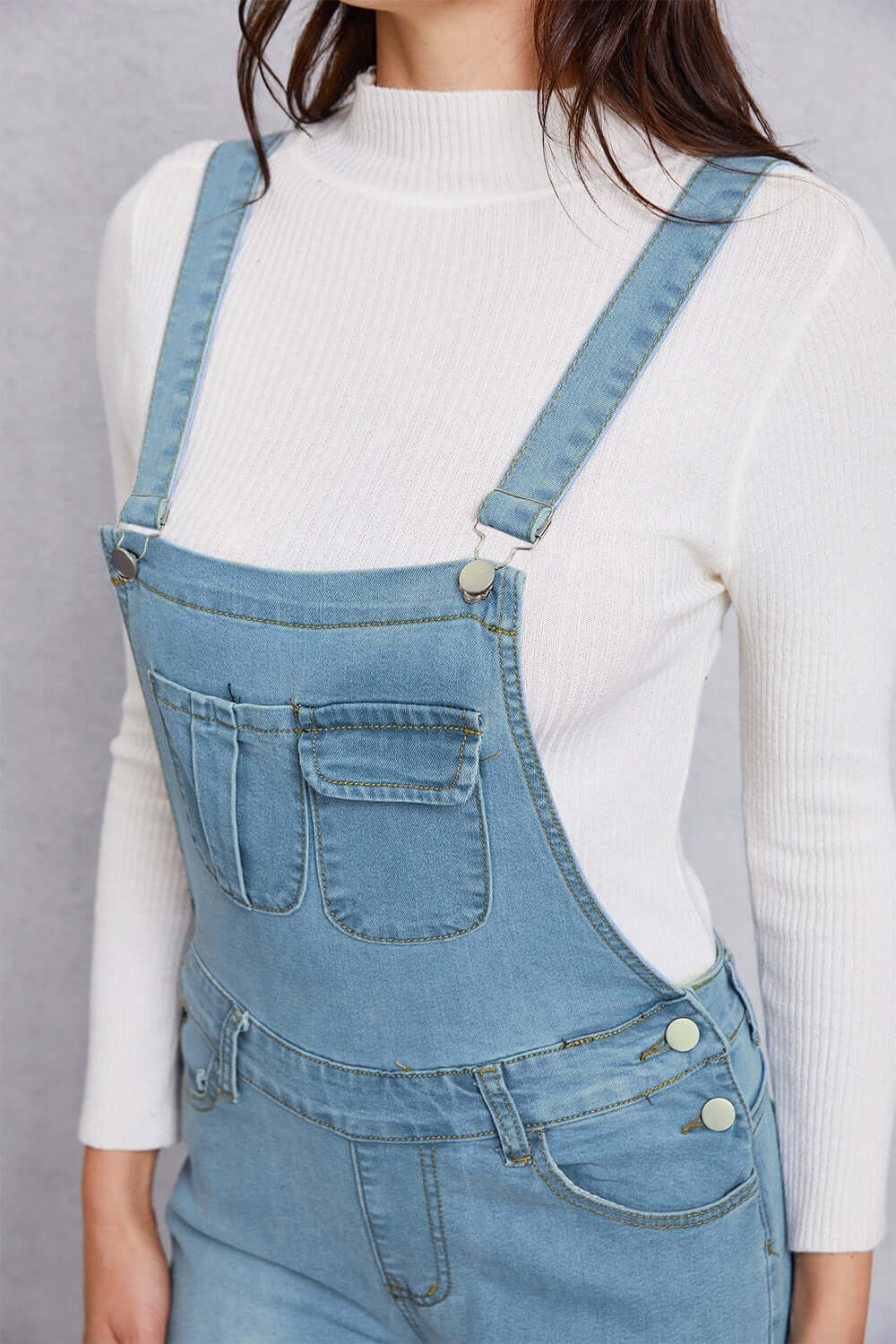 Close-up of distressed washed denim overalls with pockets, worn over a white ribbed turtleneck sweater.