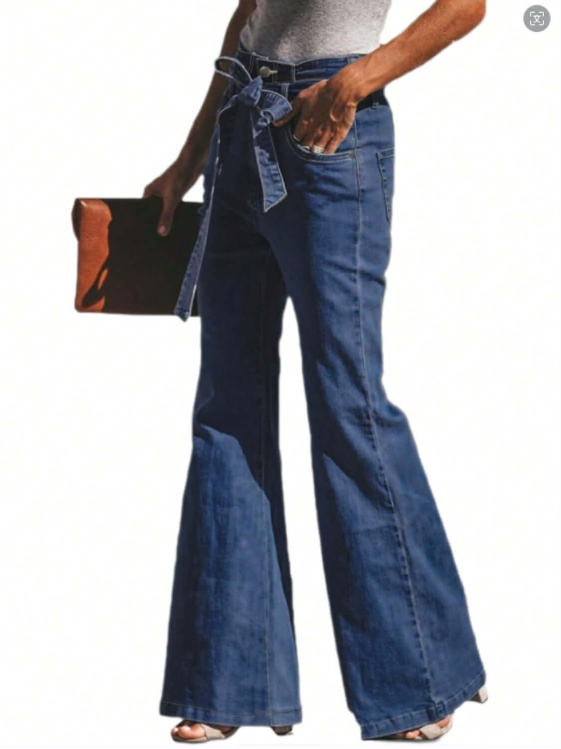 Woman wearing blue tied flare jeans with pockets and holding a brown clutch.