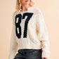 Aemi+Co cream cable knit sweater with black number 87, paired with a maroon cap and denim shorts for a casual look.