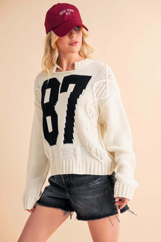 Aemi+Co cream cable knit sweater with black number 87, paired with a maroon cap and denim shorts for a casual look.