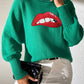 Woman wearing green Bella Road Lip Turtleneck Sweater with red lip design, paired with white pants and black clutch.