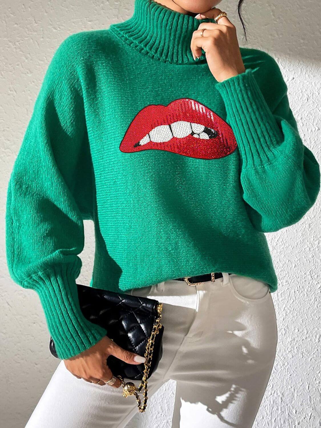 Woman wearing green Bella Road Lip Turtleneck Sweater with red lip design, paired with white pants and black clutch.