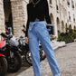 Fashionable woman in Bella Road Tied Straight Jeans with pockets, standing stylishly on a cobblestone street with motorcycles in the background.