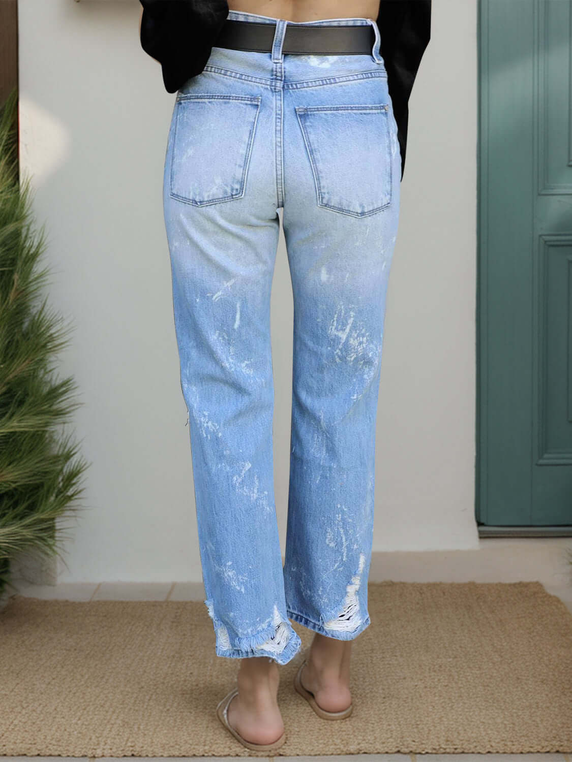 Back view of distressed straight jeans with pockets in light blue, worn by a model standing in front of a green door.