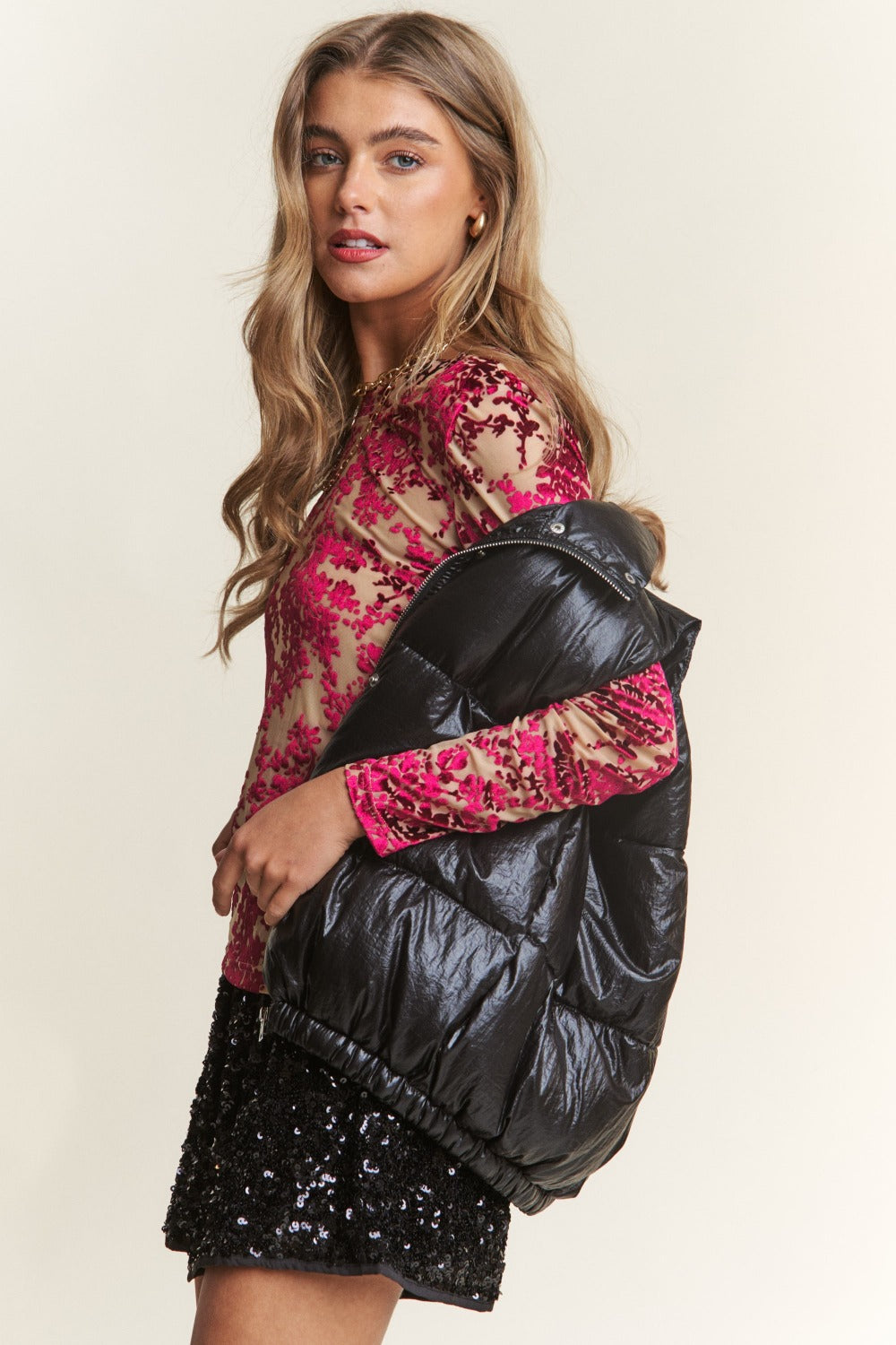 Stylish woman wearing a black shiny metallic puffer vest over a red lace top and black sequin skirt.