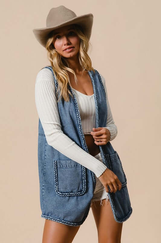 Stylish woman in a BiBi braided trim open front denim vest with pockets, paired with a hat and top, showcasing chic fashion.