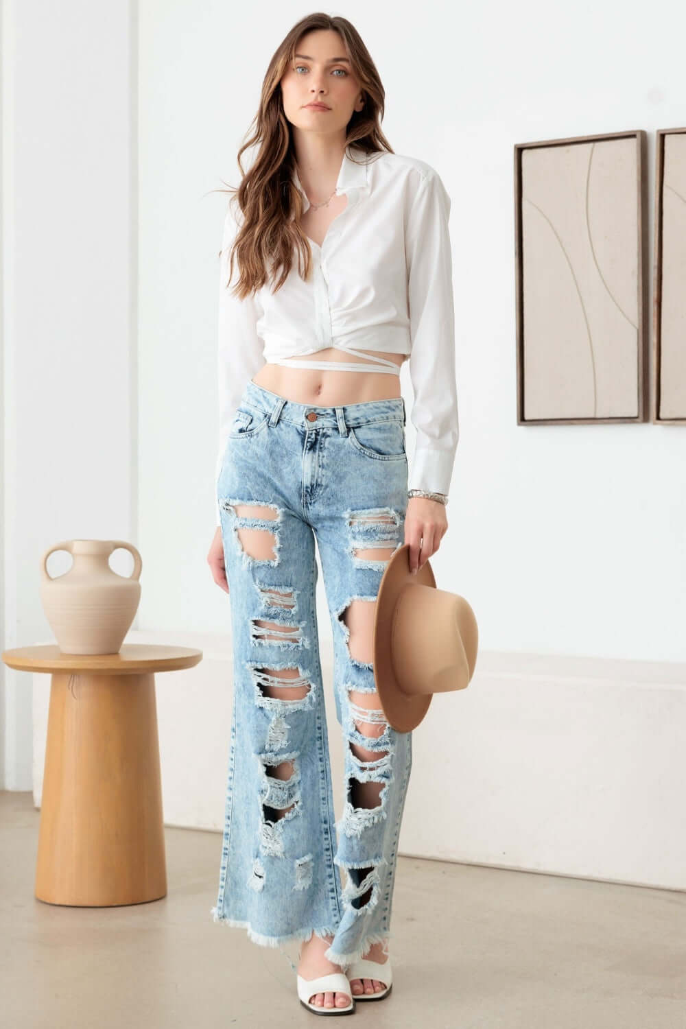 Trendy woman in distressed frayed hem flare jeans holding hat in stylish room with modern decor.