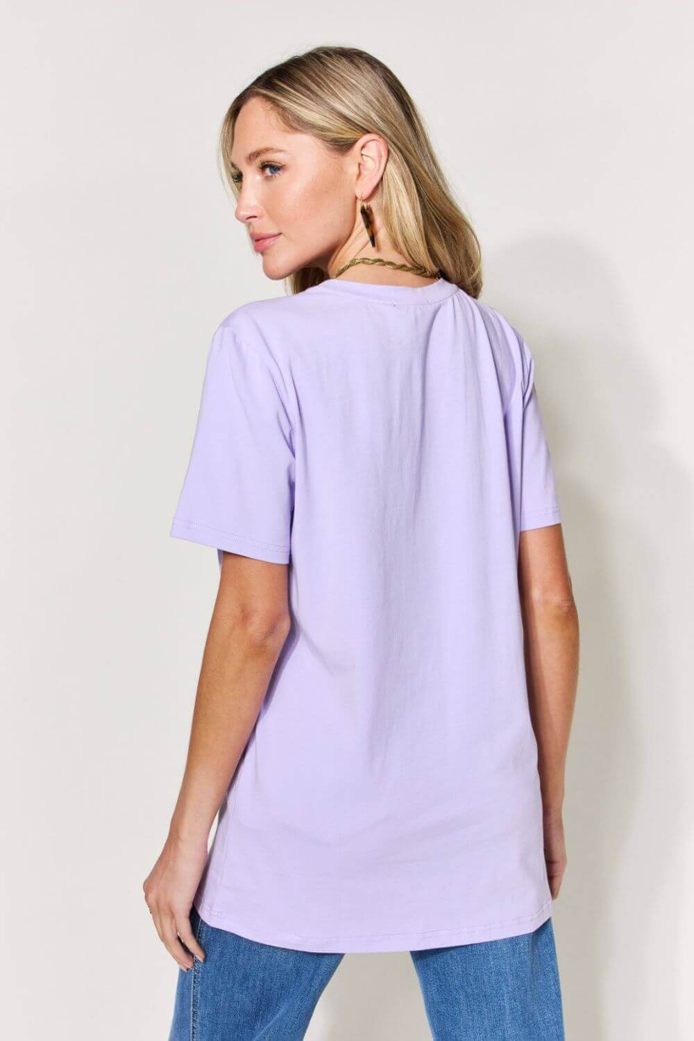 Woman wearing a lavender graphic round neck short sleeve t-shirt, slightly stretching the back, paired with blue jeans.