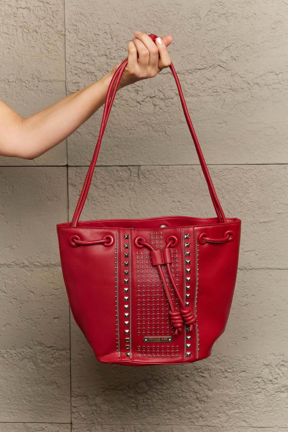 Nicole Lee USA red Amy Studded Bucket Bag with pebbled vegan leather, studded detail, and secure drawstring closure held by a hand.