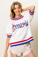 Woman wearing metallic America letter short sleeve sweater with red, white, and blue detailing, paired with distressed shorts.