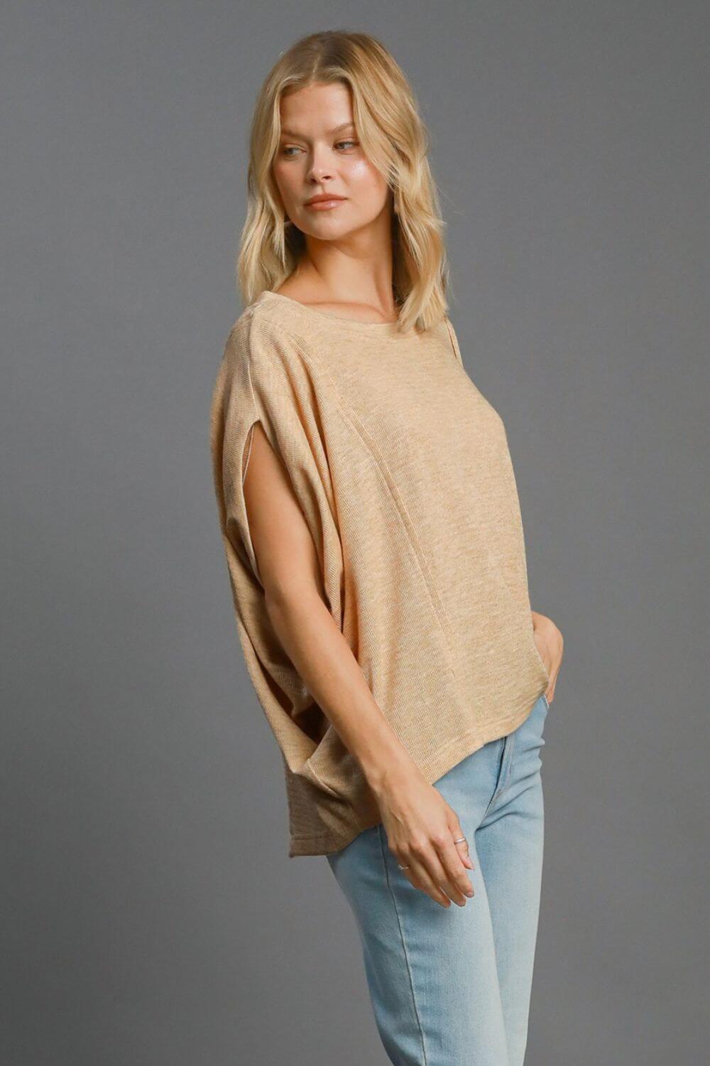 Stylish woman in a tan batwing sleeve knit top, showcasing exposed seams, paired with light blue jeans.