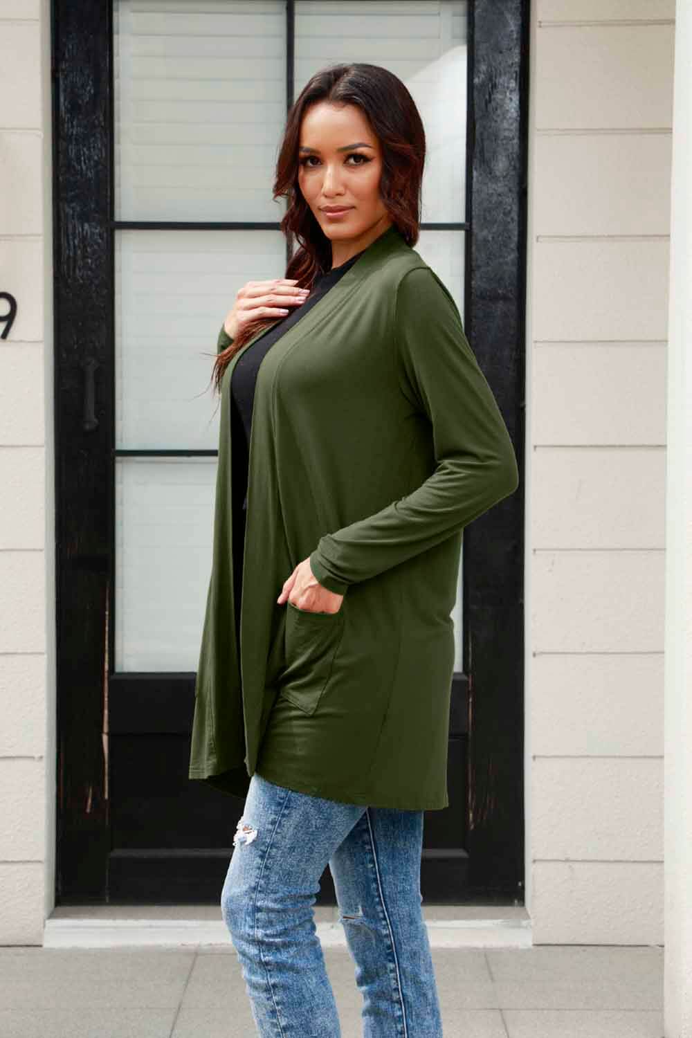 Woman wearing an open front long sleeve green cardigan with pockets over a black top and blue jeans standing in front of a door.