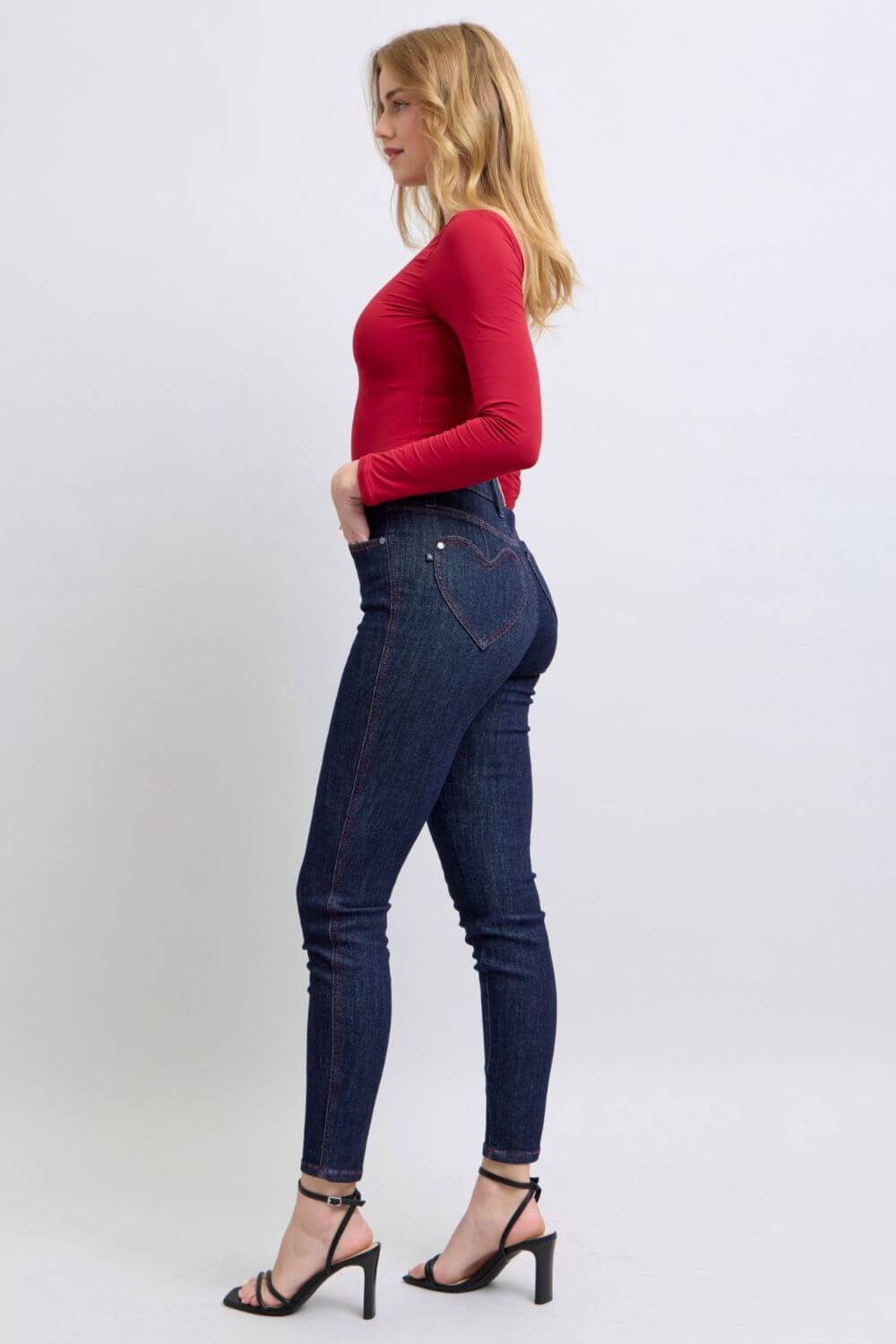 Woman wearing Judy Blue skinny jeans with heart-shaped back pockets, styled with a red top and heels, showcasing a chic silhouette.