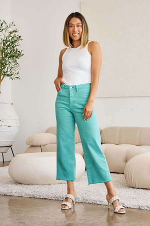 Woman wearing teal high waist raw hem jeans and white tank top, standing in stylish modern living room.