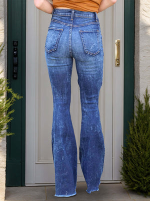 Person wearing raw hem distressed bootcut jeans in denim color, standing in front of a door.