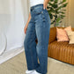 Woman wearing RFM full size high rise tummy control wide leg jeans in blue, standing in stylish modern living room.