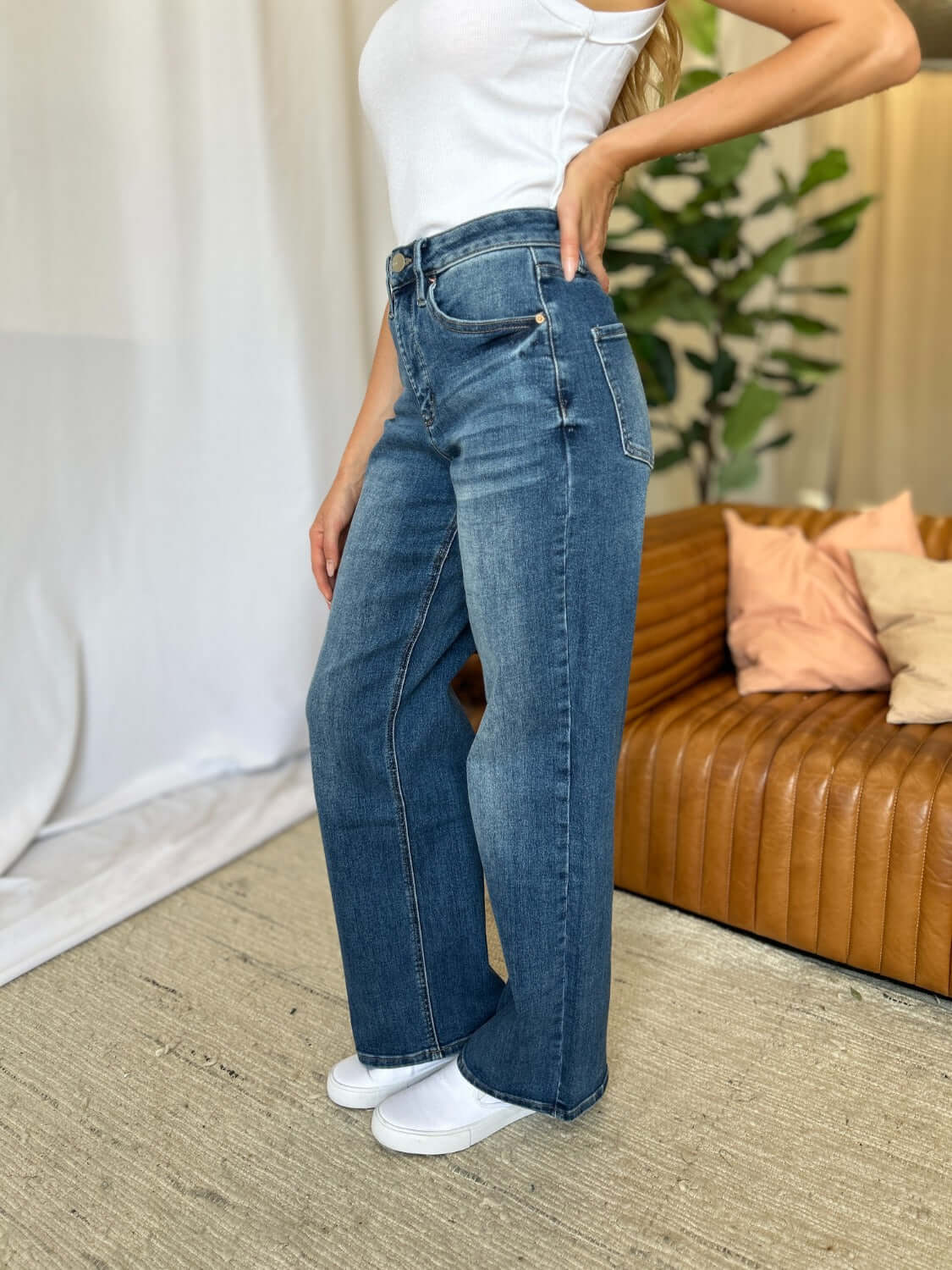 Woman wearing RFM full size high rise tummy control wide leg jeans in blue, standing in stylish modern living room.