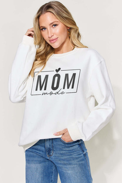 Woman wearing white letter graphic long sleeve sweatshirt with "Mom Mode" text, paired with blue jeans.