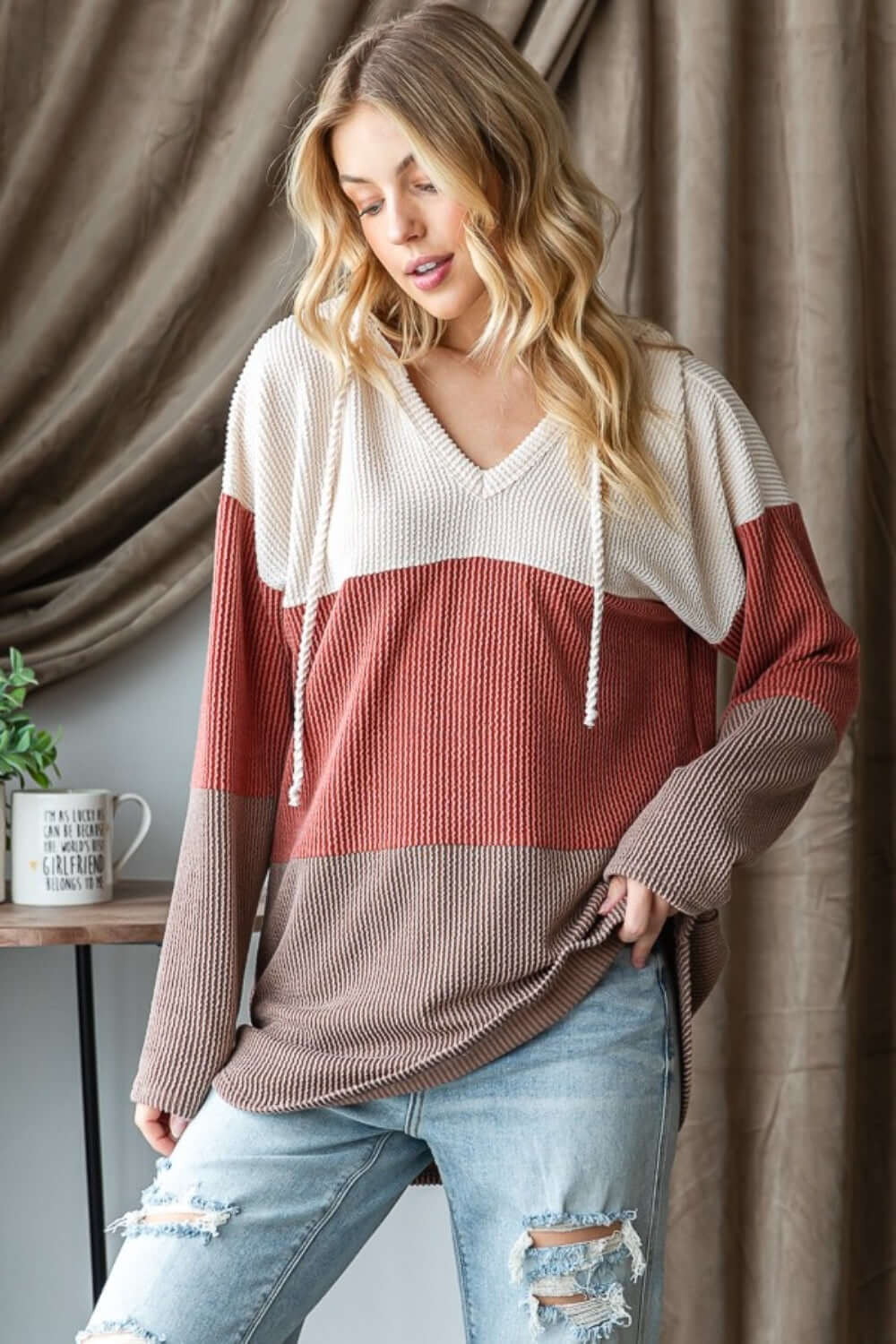 Woman wearing Color Block Long Sleeve Hoodie with trendy design and ripped jeans, standing indoors against a curtain background.