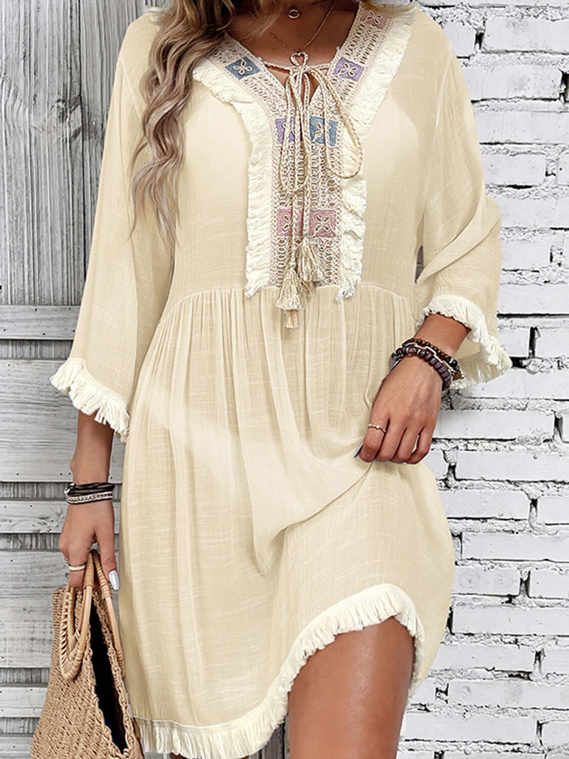 Woman wearing a beige sheer fringe tie neck three-quarter sleeve cover-up with embroidered detailing, holding a woven bag.