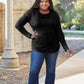 Woman wearing a black round neck long sleeve t-shirt and blue jeans, smiling outdoors near an architectural structure and greenery.