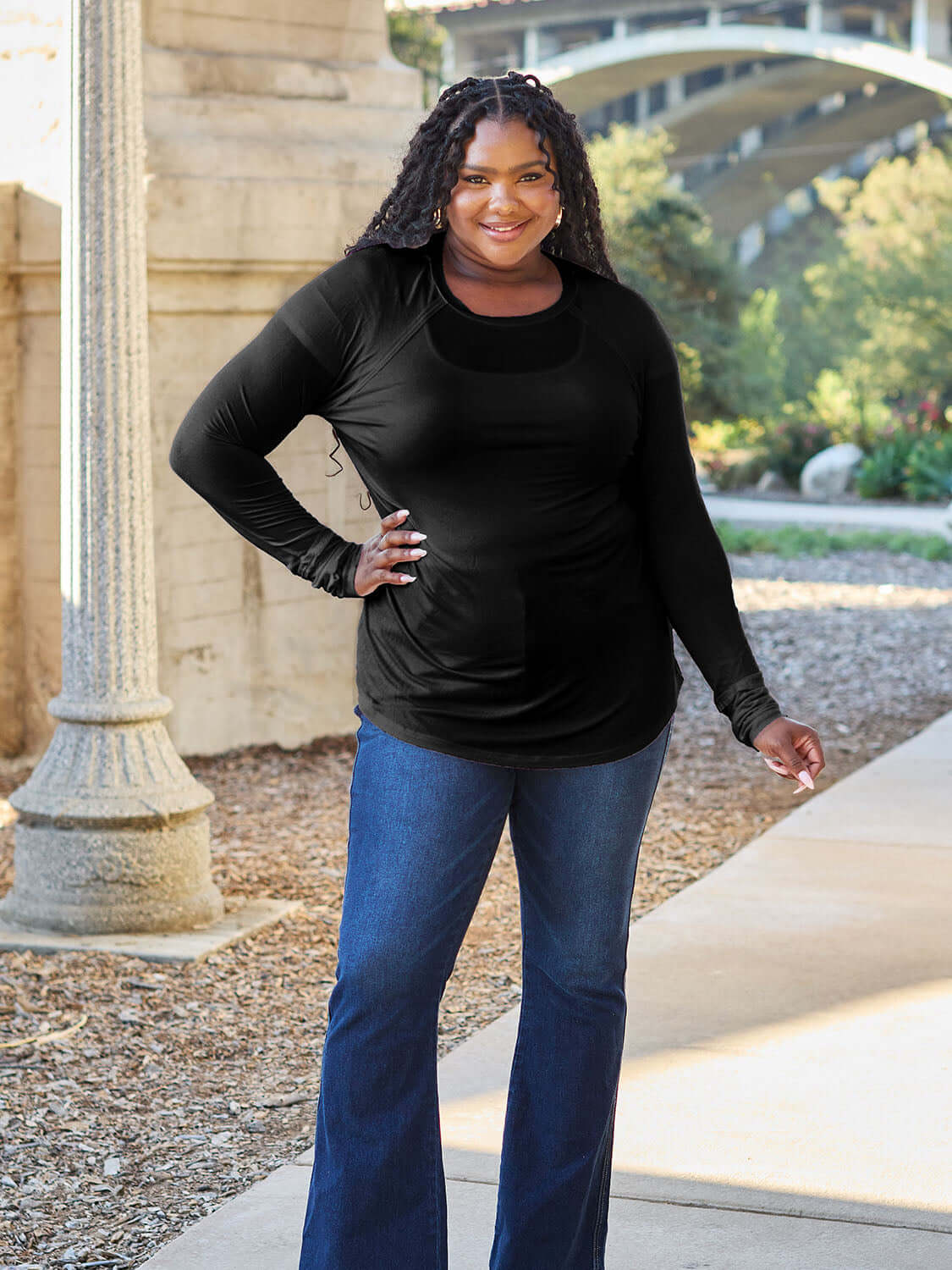 Woman wearing a black round neck long sleeve t-shirt and blue jeans, smiling outdoors near an architectural structure and greenery.