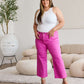 Woman wearing pink high waist raw hem jeans and white tank top, standing in a modern living room setting.