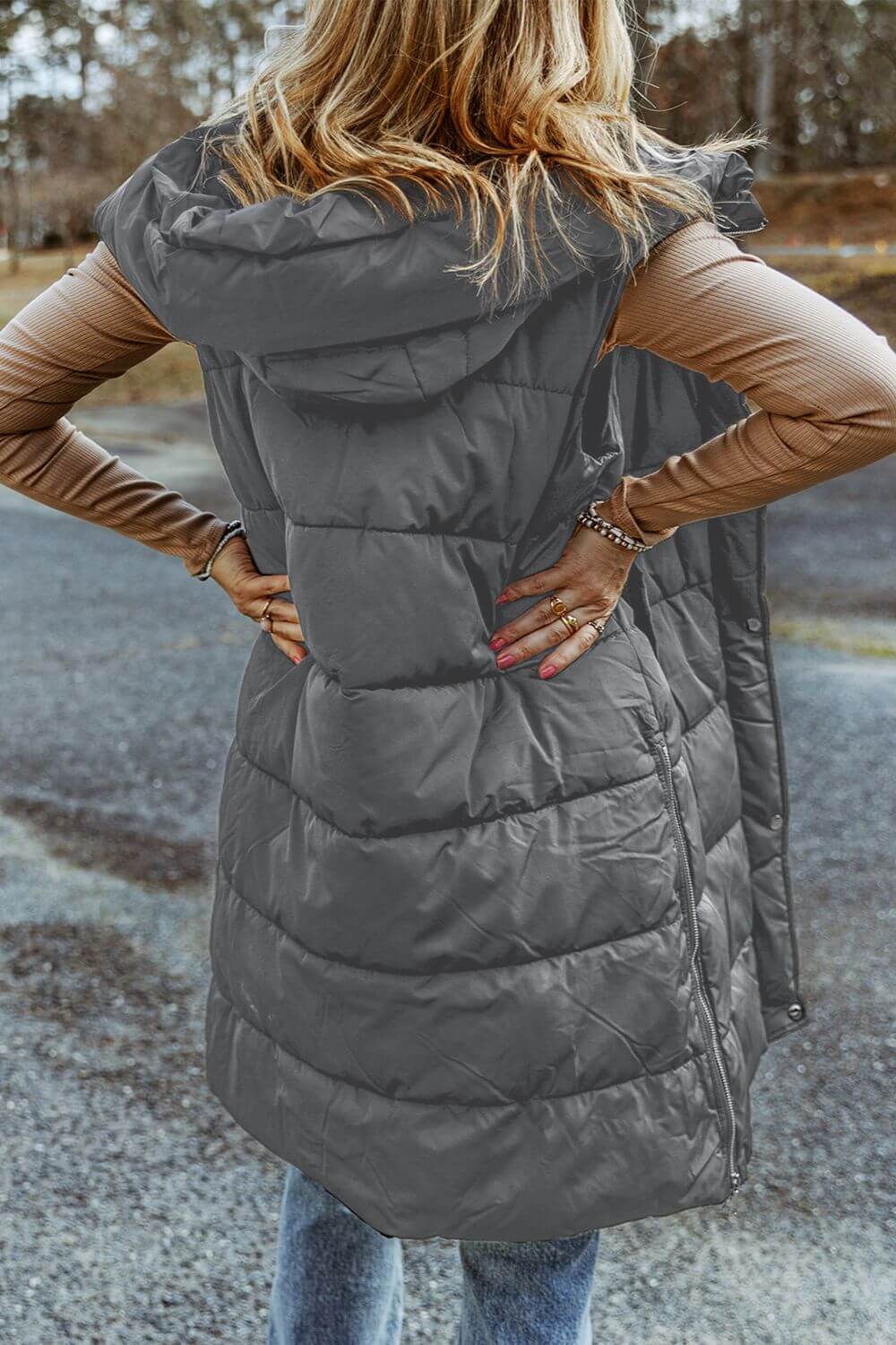 Woman in Bella Road Longline Hooded Sleeveless Puffer Vest stands outdoors with hands on hips, showcasing its stylish comfort.