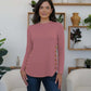 Woman wearing FAM-FAM dusty pink round neck long sleeve t-shirt with decorative buttons, standing in stylish living room.