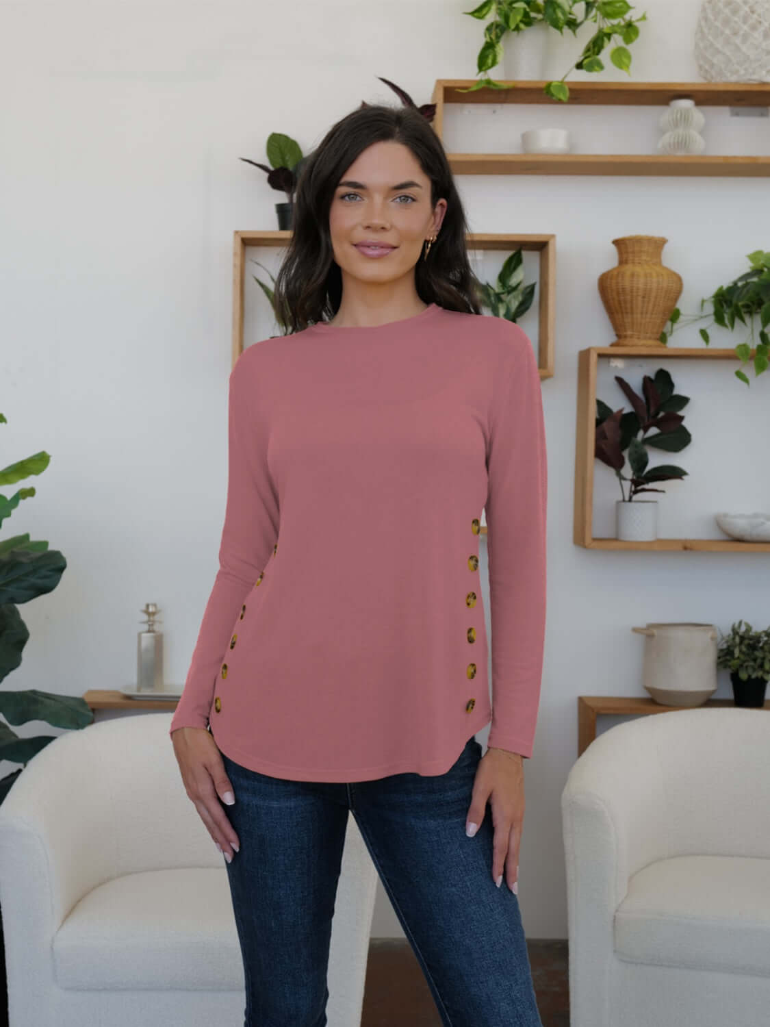 Woman wearing FAM-FAM dusty pink round neck long sleeve t-shirt with decorative buttons, standing in stylish living room.