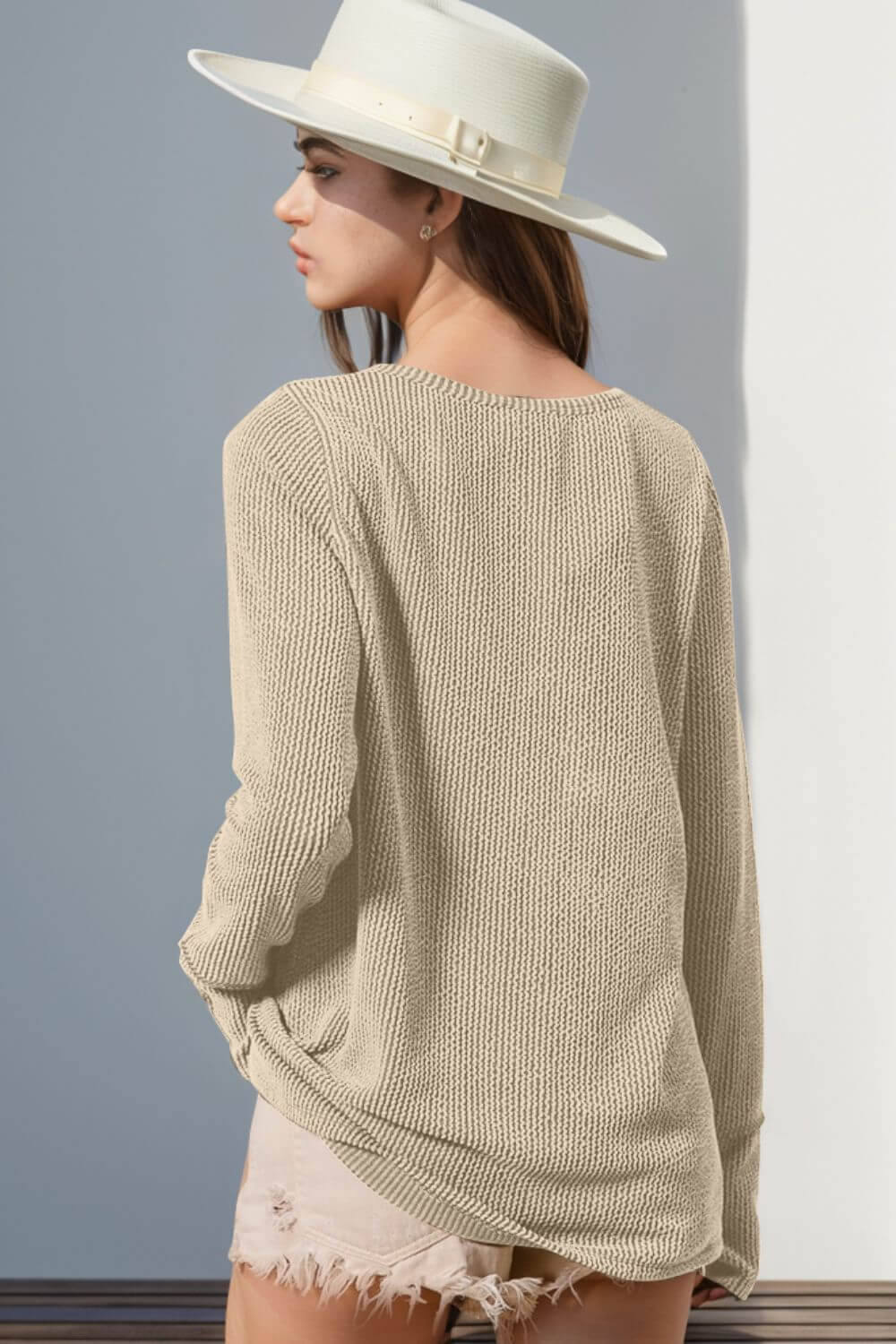 Woman wearing beige knit long sleeve sweater with thumbholes, paired with white hat and distressed shorts.