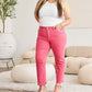 Woman wearing pink high-waist raw hem jeans with belly control, paired with a white top, standing confidently in a stylish room. RFM Jeans