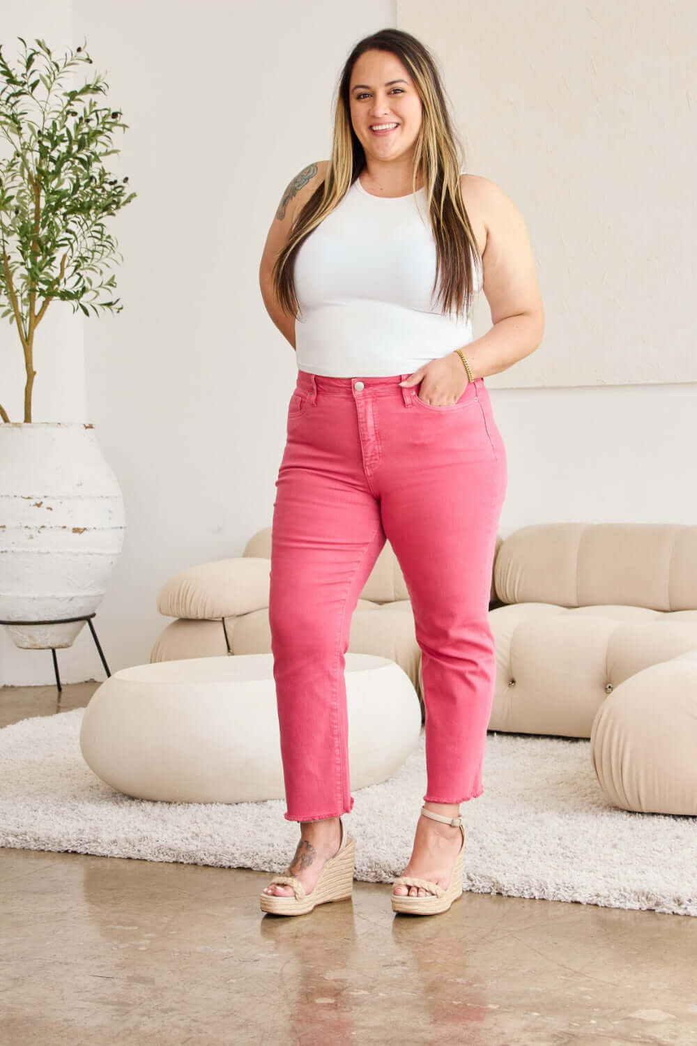 Woman wearing pink high-waist raw hem jeans with belly control, paired with a white top, standing confidently in a stylish room. RFM Jeans