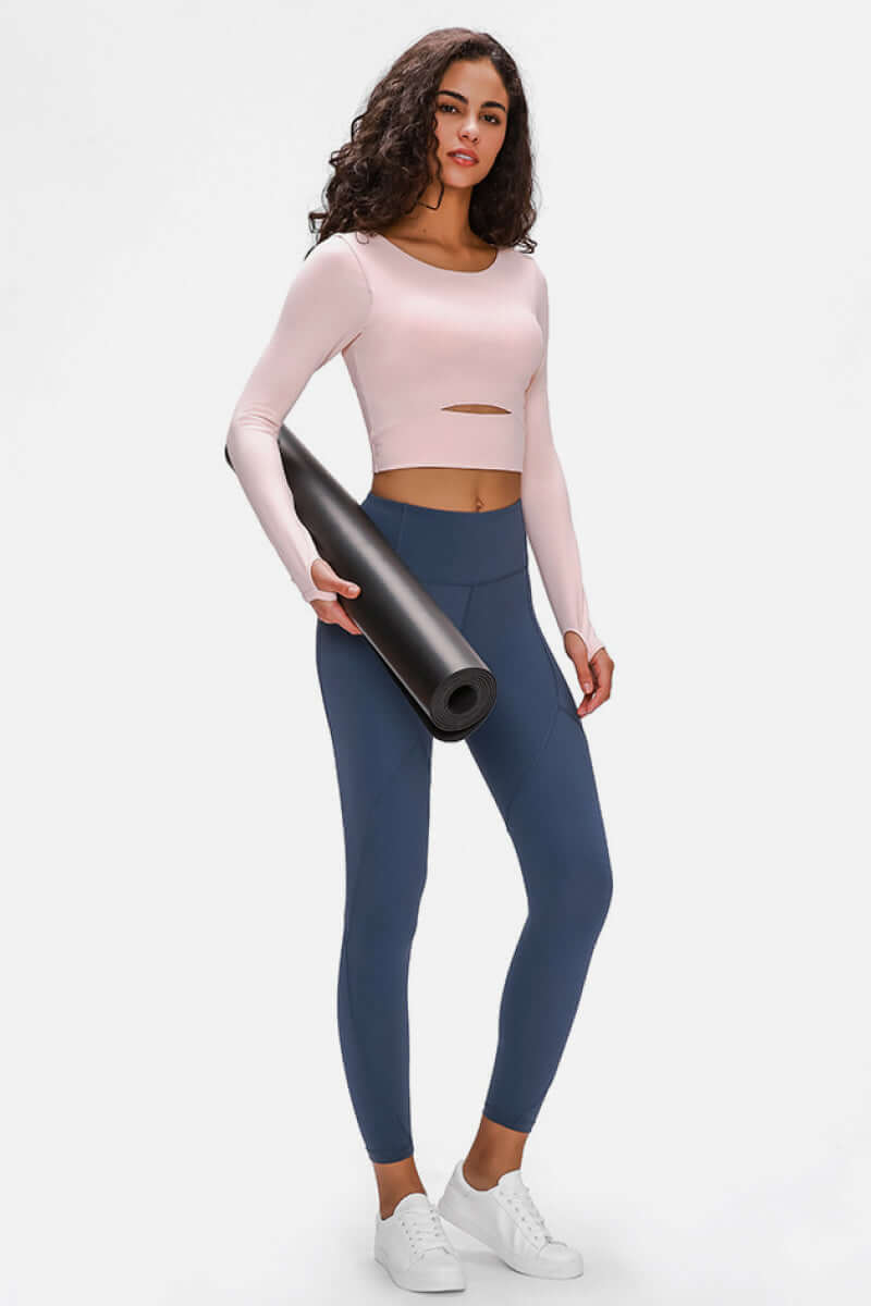 Woman in a pink long-sleeved cropped top and blue leggings, holding a yoga mat, ready for a workout.