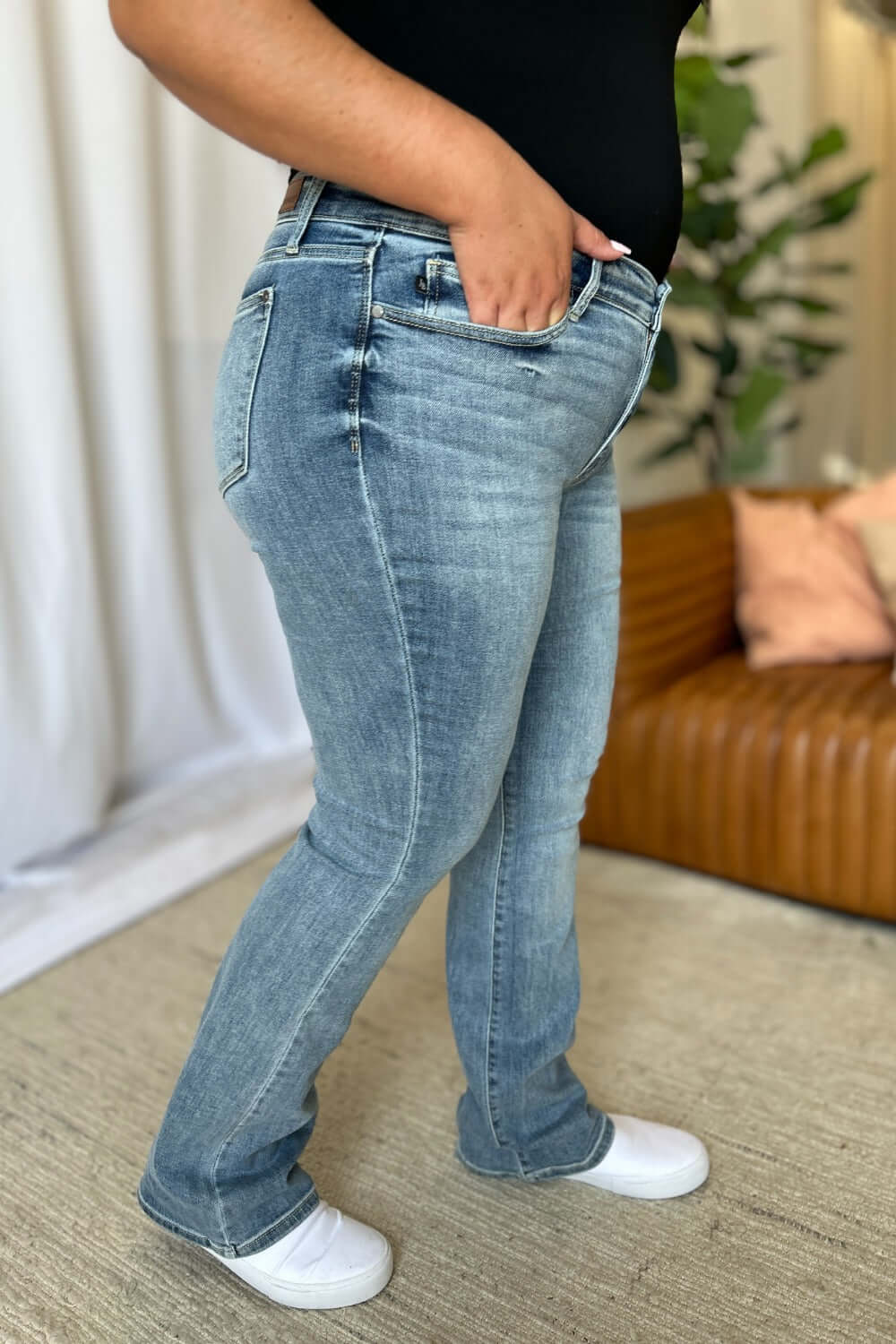 Woman modeling Judy Blue medium rise bootcut jeans with white sneakers in a living room setting.