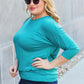 Woman wearing a turquoise round neck batwing sleeve top, paired with dark blue jeans, posing outdoors.