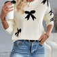 Cozy white sweater with playful black bow graphics, round neck, and long sleeves, worn with casual blue jeans.