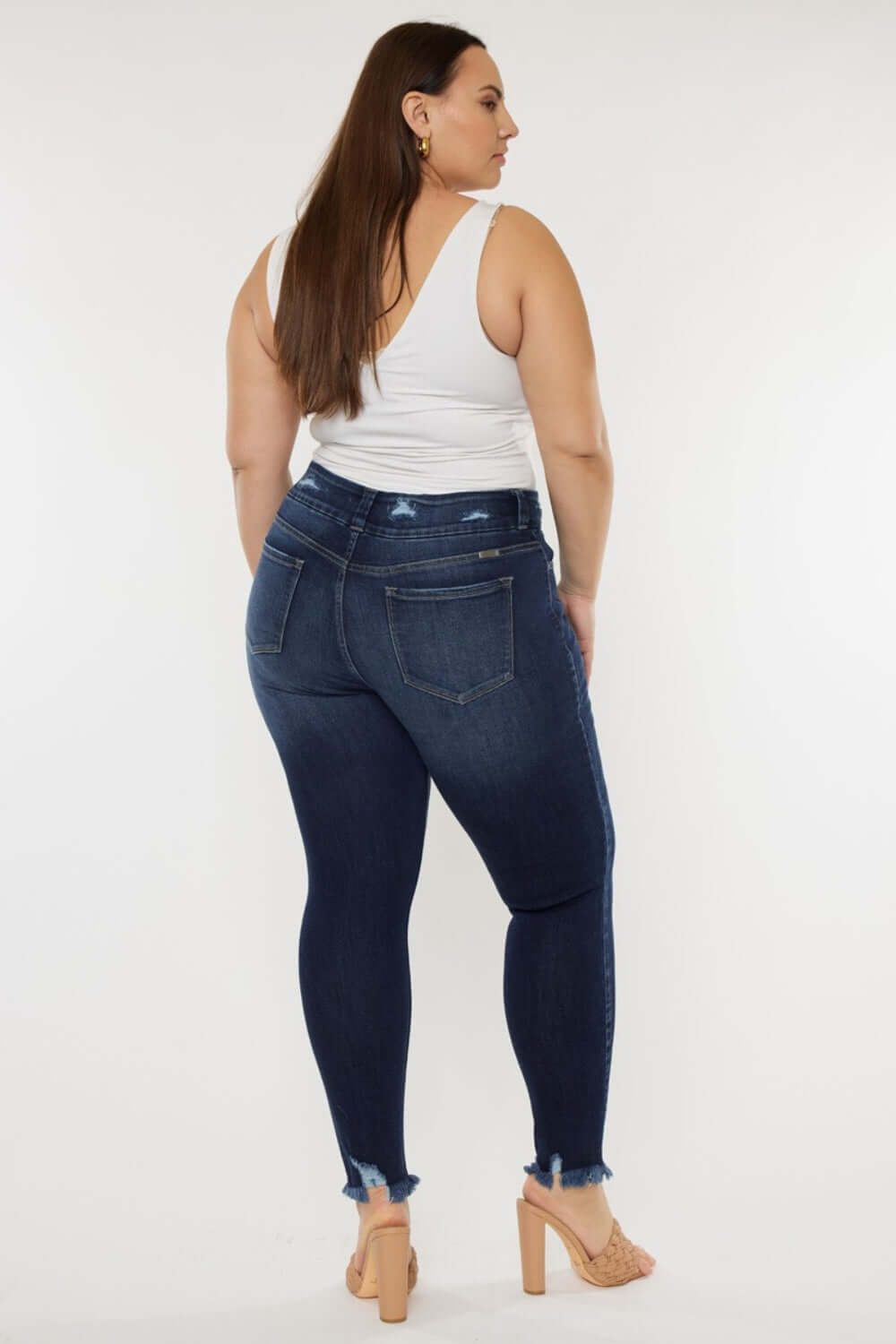 Woman modeling high waisted raw hem jeans in dark blue with a white tank top and heels, showing back view